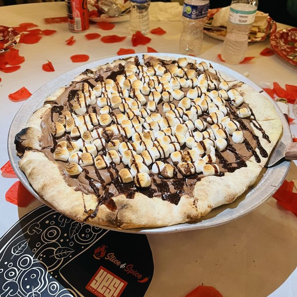 A dessert pizza from Slice & Spice, topped with melted marshmallows and chocolate drizzle on a table with rose petals.